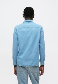 Gaiši zila džinsa krekls ar apkakli, garām piedurknēm un pogu aprocēm. Raksturo gluda tekstūra un taisna apakša.