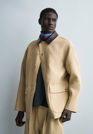 Άνδρας που φοράει μπεζ σακάκι και matching παντελόνι με σκούρο γιακά και πολύχρωμο ριγέ κασκόλ, κοιτάζοντας προς τα πλάγια με φόντο μονόχρωμο φόντο.