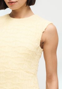 Γυναίκα με κοντά σκουρόχρωμα μαλλιά που φοράει ένα αμάνικο, με υφή, ανοιχτό κίτρινο τοπ μπροστά σε απλό ανοιχτό φόντο.