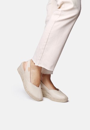 Kāji valkā bēša krāsas wedge espadrilles kopā ar gaismas toņa saīsinātām biksēm uz baltas vienkrāsainas aizmugures.