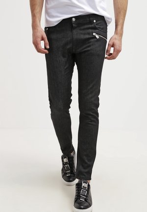 Man draagt strakke zwarte jeans met een diagonale ritszak en zwarte sneakers met witte zolen, lopend naar voren.