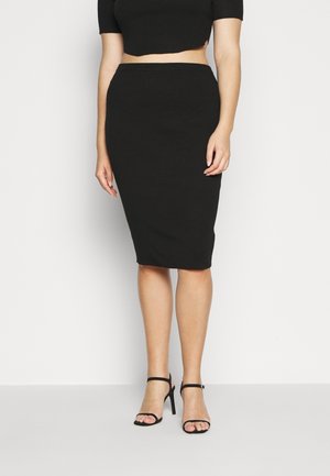 Vrouw die een zwarte hooggetailleerde kokerrok draagt met een bijpassende cropped top en zwarte sandalen met hoge hakken, staand tegen een witte achtergrond.