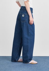 Vaqueros de mezclilla azul de pierna ancha con una cintura alta, que cuentan con un bolsillo trasero con una etiqueta de logo blanca. La tela parece resistente y de textura suave.