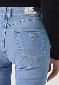 Jeans de mezclilla azul claro con un diseño ajustado. Cuenta con un bolsillo trasero triangular con el logo "GUESS". Las costuras son visibles a lo largo de las uniones.
