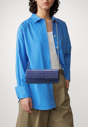 Personne portant une chemise rayée bleu et blanc, un pantalon beige, tenant une pochette rectangulaire bleue perlée avec une bandoulière en chaîne argentée.