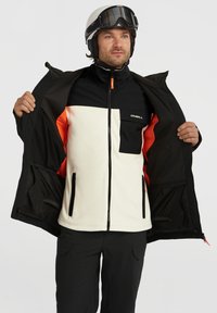 Zwarte en crèmekleurige skijas met een oranje voering. Voorzien van ritszakken, hoge kraag en een logo op de borst. Gladde textuur.