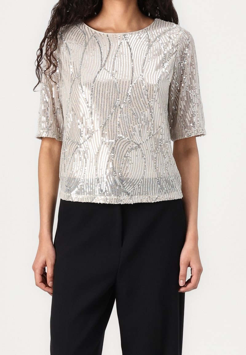 Femme portant un haut court à sequins argentés avec des motifs abstraits et un pantalon noir sur un fond uni.