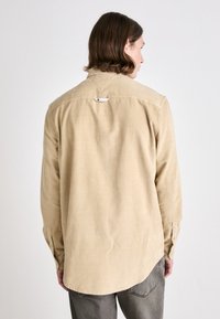 Camicia di velluto a coste beige con maniche lunghe, colletto a punta e polsini con bottoni. Presenta un piccolo etichetta del marchio nella parte posteriore del girocollo.