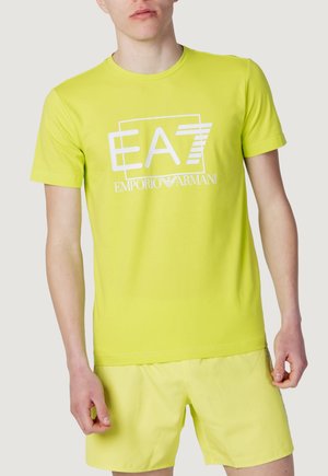 Mann trägt ein hellgelbes EA7 Emporio Armani T-Shirt und passende Shorts, steht vor einem einfachen hellen Hintergrund.