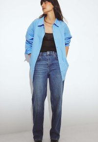 Šviesiai mėlyna marškinėliai su pasuktomis rankovėmis, uždėti ant juodos viršūnės, derinami su aukštu liemeniu, laisvai krentančiais mėlynais džinsais.