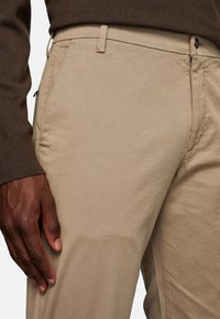 Beige cotton trousers with a smooth texture, featuring side pockets, a zipper, and a single button closure. Close-up view.