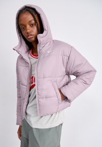 Gaiši violeta pūkains mētelis ar kapuci, ar rāvējslēdzēju priekšā un sānu kabatām; uz krūtīm ir neliela Nike logo. Mīksta, iešūta tekstūra.