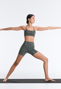 Femme en tenue de sport verte à motifs floraux, tenant les bras et jambes tendus en posture de guerrier de yoga sur un tapis noir sur fond blanc.