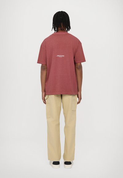 Camiseta de algodón color burdeos con mangas cortas y cuello redondo, con un diseño de texto blanco en la parte posterior, combinada con pantalones cargo beige.