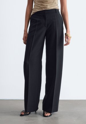 Femme portant un pantalon noir taille haute à jambes larges, un haut beige, des talons ouverts noirs et des bracelets dorés aux deux poignets.