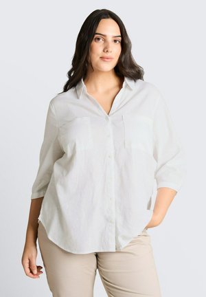 Frau mit langen dunklen Haaren, die ein weißes Hemd mit Knöpfen und beige Hose trägt, steht mit einer Hand in der Tasche vor einfarbigem Hintergrund.