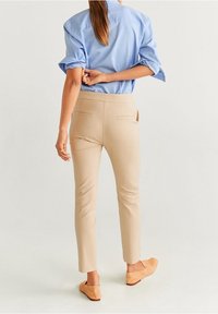 Kobieta w beżowych, dopasowanych spodniach, niebieskiej, podwiniętej bluzie z długim rękawem i jasnobrązowych butach wsuwanych, stojąca tyłem.