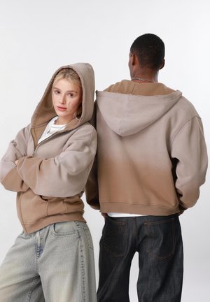 Nuori nainen ja mies pukeutuneina beigeihin hupullisiin takkeihin, nainen katsoo eteenpäin käsivarret ristissä, mies katsoo taaksepäin, molemmat pukeutuneet rentoihin farkkuihin.