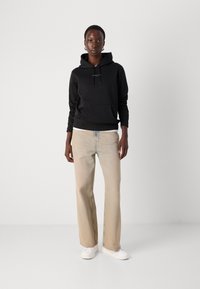 Sudadera negra con cordones y bolsillo frontal, combinada con jeans rectos de un beige claro. El modelo lleva zapatillas blancas.