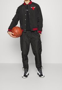 Svart bomberjacka med röda ränder och logga, svarta cargo-byxor och svartvita sneakers, håller en basketboll.