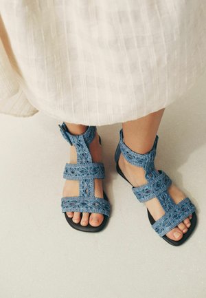 Füße, die blaue gehäkelte Sandalen mit schwarzen Sohlen tragen, stehen auf einem hellen Boden, teilweise bedeckt von einem weißen strukturierten Rock.