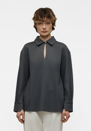 Rövid hullámos hajú személy sötétszürke, gombos galléros hosszú ujjú felsőben és fehér nadrágban, egyszínű háttér előtt állva.