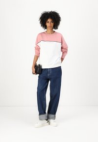 Hvit og lys rosa genser med "GUESS JEANS"-logo, sammen med løse blå jeans og hvite joggesko. Svart håndveske i hånden.