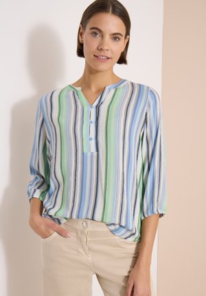 Donna che indossa una camicetta larga a righe verticali blu, verdi e bianche e pantaloni beige, in piedi con una mano in tasca.