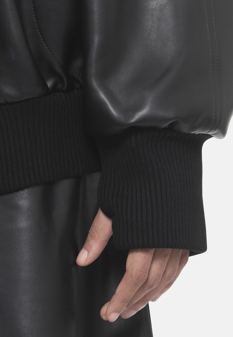 Giacca in pelle nera con polsini in maglia nera a costine, caratterizzata da una texture liscia e una vestibilità rilassata, che enfatizza elementi di design contemporaneo.