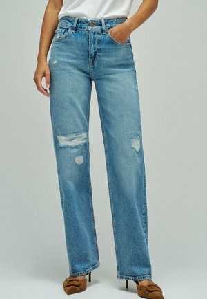 Mujer con vaqueros de tiro alto de pierna recta azul claro con parches desgastados, top blanco y zapatos de tacón alto marrones.