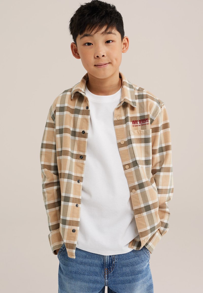 Garçon aux cheveux noirs courts portant une chemise à carreaux beige boutonnée sur un t-shirt blanc et un jean bleu, debout, les mains dans les poches.