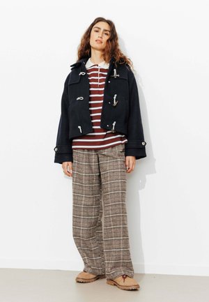Manteau duffle court marine avec des fermetures à bascule, chemise rayée bordeaux et blanche en dessous, pantalon à carreaux à jambes larges, et chaussures beiges.