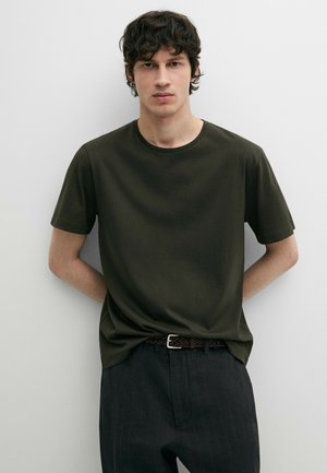 Jonge man met donker krullend haar, gekleed in een donkergroen T-shirt met korte mouwen en zwarte krijtstreepbroek, staand tegen een eenvoudige lichte muur.