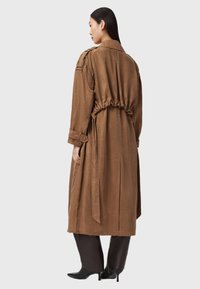 Hnědý trench coat s dlouhým rukávem a stahovacím pasem, vyrobený z hladké tkaniny. Obsahuje ramenní epauletky a pásek vázaný vzadu.