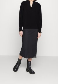 Musta pitkähihaisuus V-pääntie, yhdistetty mustaan satiinisiin midihameeseen ja tukevasiin mustiin nahkasaappaisiin. Sujuva pinta ja minimalistinen muotoilu.