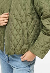 Asmuo dėvi žalią quiltuotą striukę su priekiu užtrauktuku ir šoninėmis kišenėmis, derinančias prie mėlynų džinsinių kelnių.