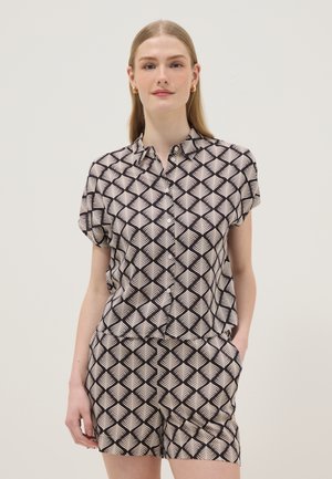 Giovane donna che indossa una maglietta a maniche corte e pantaloncini coordinati con motivo geometrico nero e beige, in piedi con una mano in tasca.