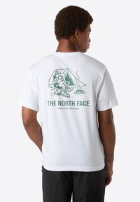 Muž s bílým tričkem, na kterém je zelená scéna t�áboráku a text "THE NORTH FACE Never Stop Exploring" na zadní straně, otočený zády k fotoaparátu.