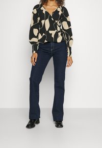 Top avvolgente con motivo nero e beige, maniche lunghe, abbinato a jeans a zampa d'elefante blu e stivali neri con tacco. Tessuto liscio.