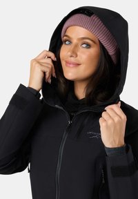 Sort vandtæt jakke med lynlås foran og hætte. Har et synligt mærkelogo, velcro-detaljer og en struktureret strikket lyserød hue.