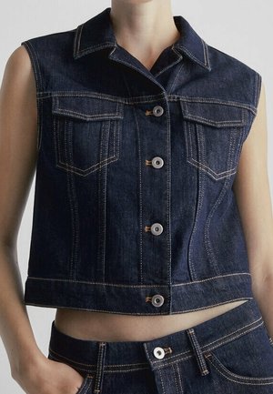 Mouwloos donker denimvest met knoopsluiting aan de voorkant, borstflappzakken en contrasterend stiksel, gedragen over bijpassende donkere denim jeans.