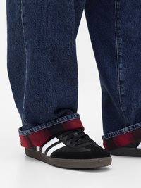 Baskets noires avec des rayures blanches et un daim texturé, associées à un jean en denim bleu foncé avec un revers à carreaux rouges.