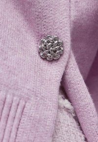 Fioletowy wełniany sweter o teksturowanym wykończeniu, z metaliczną srebrną broszką w kształcie kwiatu jako akcentem. Zbliżenie na tkaninę i detale.