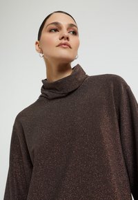 Brauner, glitzernder Rollkragenpullover mit lässigem Schnitt und umgeschlagenem Kragen; der Stoff hat einen dezenten Schimmer und eine glatte Textur.