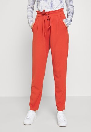 Person iført røde, højtaljede, tilspidsede bukser med lommer og snøre i taljen, kombineret med hvide sneakers og en langærmet mønstret top.