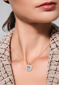 Złoty naszyjnik z łańcuszka z kwadratowym, jasnoniebieskim kamieniem otoczonym halo małych, przezroczystych kamieni, noszony na tle fakturowanej beżowej tkaniny.