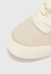 Beige velúr sneaker, texturált cipőfűzővel és puha, világos krémszínű gumitalppal. Kerek orr és kontrasztos varrási részletek.