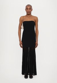 Black strapless dress with a fitted bodice and pleated sheer skirt. Model holds a small light-colored bag. Flat black shoes complete the look.