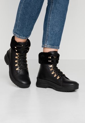 Sorte læderankelstøvler med guldøjer, sorte snørebånd og pelskant, båret med blå jeans på en hvid gulv.