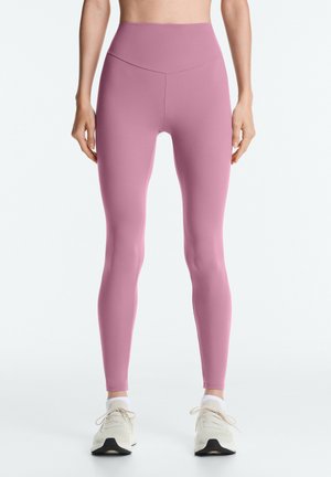 Žena oblečená vo vysokých omotaných ružových legínach a bielych teniskách, stojaca pred jednoduchým svetlým pozadím.